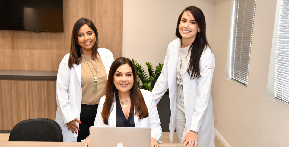 Três mulheres de jaleco estão em pé ao redor de uma mesa com um laptop.